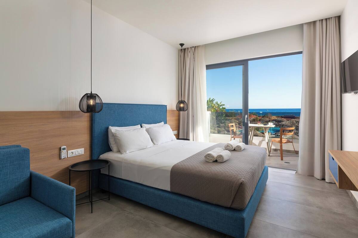A modern hotel room with a large bed, a blue sofa, and a balcony with a view of the sea.