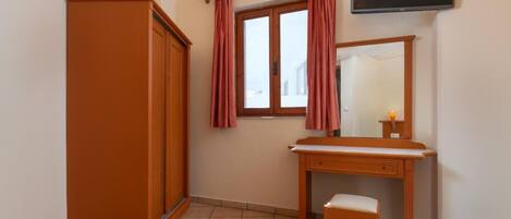 A bedroom with a bed, a wooden wardrobe, a desk with a mirror, and a window with curtains.