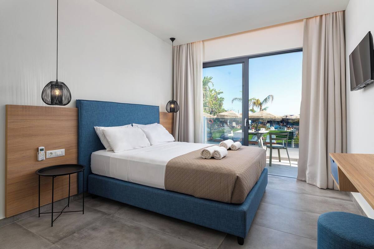 A modern hotel room with a large bed, a small table, a TV, and a balcony with a view of palm trees.