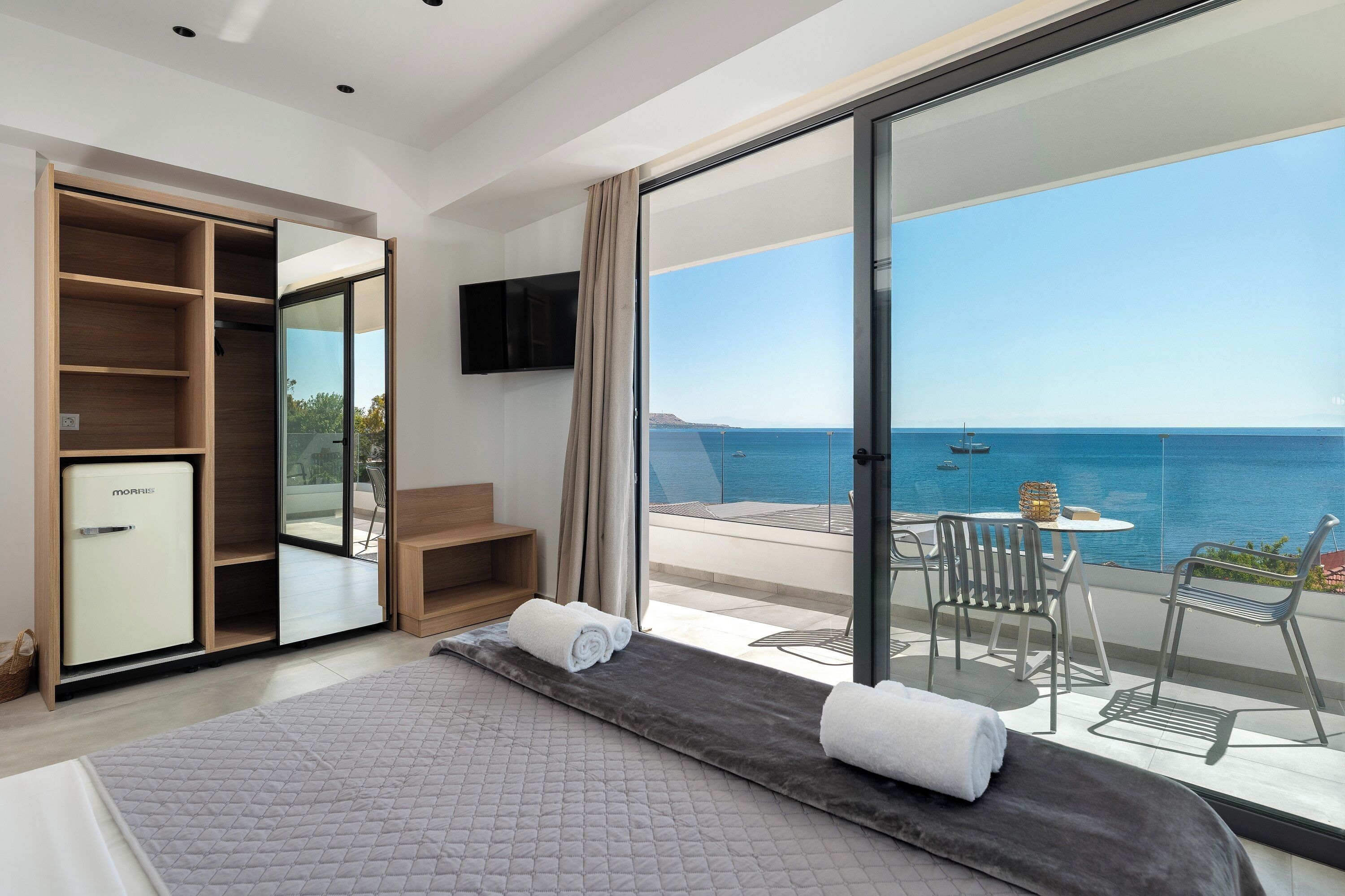 Un dormitorio moderno con una cama grande, refrigerador integrado, televisión y puerta corrediza de vidrio que da a un balcón con vista al mar.