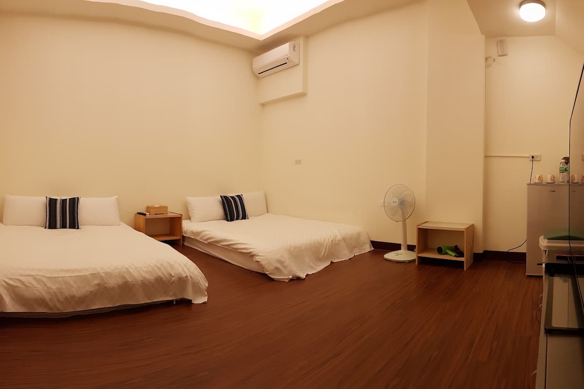 A hotel room with two beds, a standing fan, a small wooden shelf, and a TV.