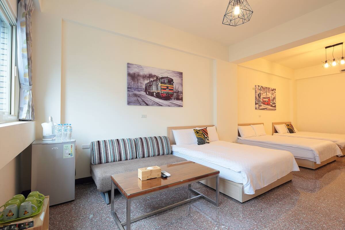 A hotel room with a bed, a sofa, a small table, a refrigerator, and artwork on the walls.