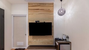 A modern living room with a flat-screen TV mounted on a wooden panel wall, a small side table with a kettle and cups, and a compact refrigerator. - Happiness Yes Inn (Luodong)