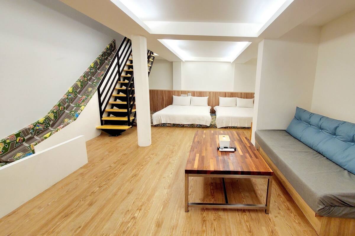 A modern hotel room with a wooden floor, a sofa, a bed with white bedding, a small table with a remote control, and a staircase with patterned carpet.