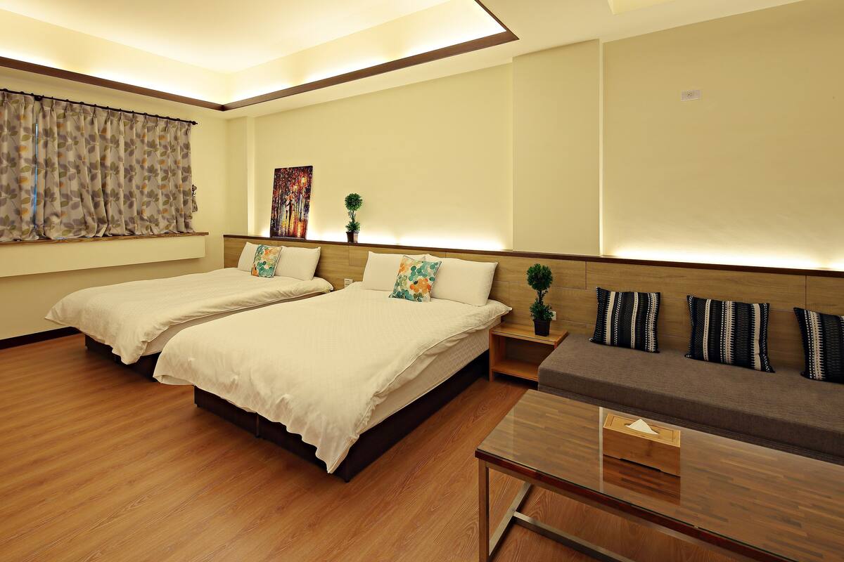 A hotel room with two beds, a sofa, a small table, and a wall-mounted TV.