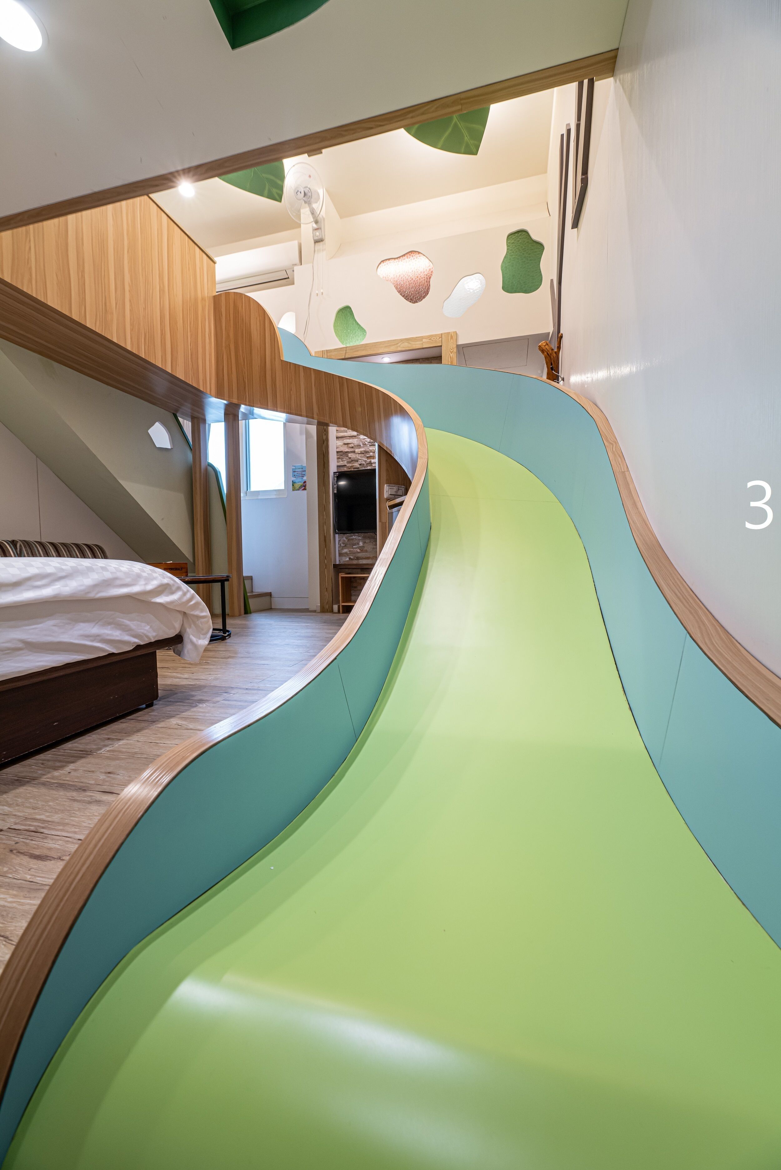 Un intérieur moderne avec un toboggan vert incurvé, un escalier en bois et un lit doté d’une tête de lit blanche.