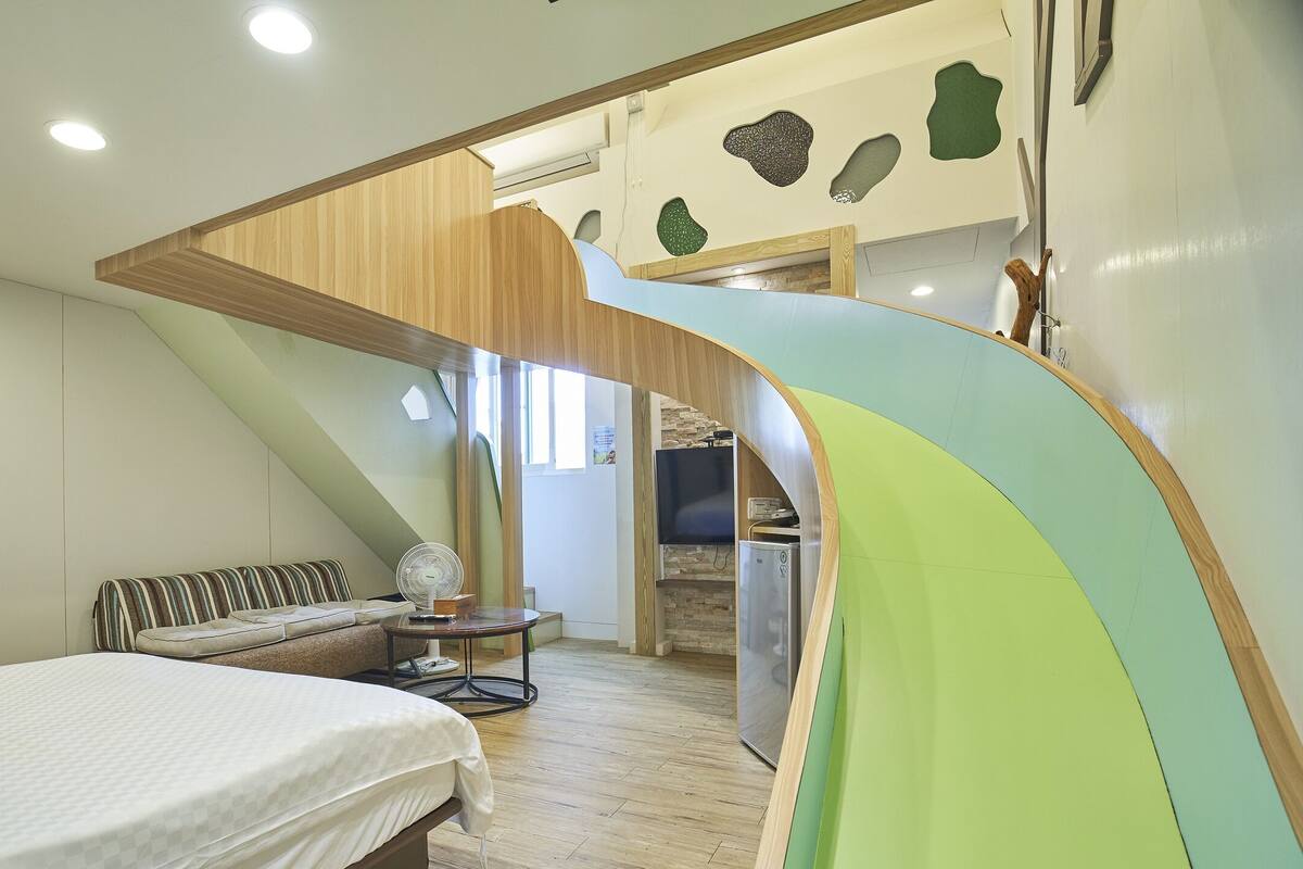 A bedroom with a bed, a small table, a fan, and a kitchenette area.