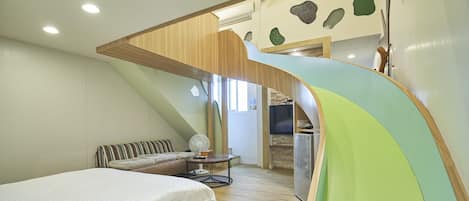 A bedroom with a bed, a small table, a fan, and a kitchenette area.