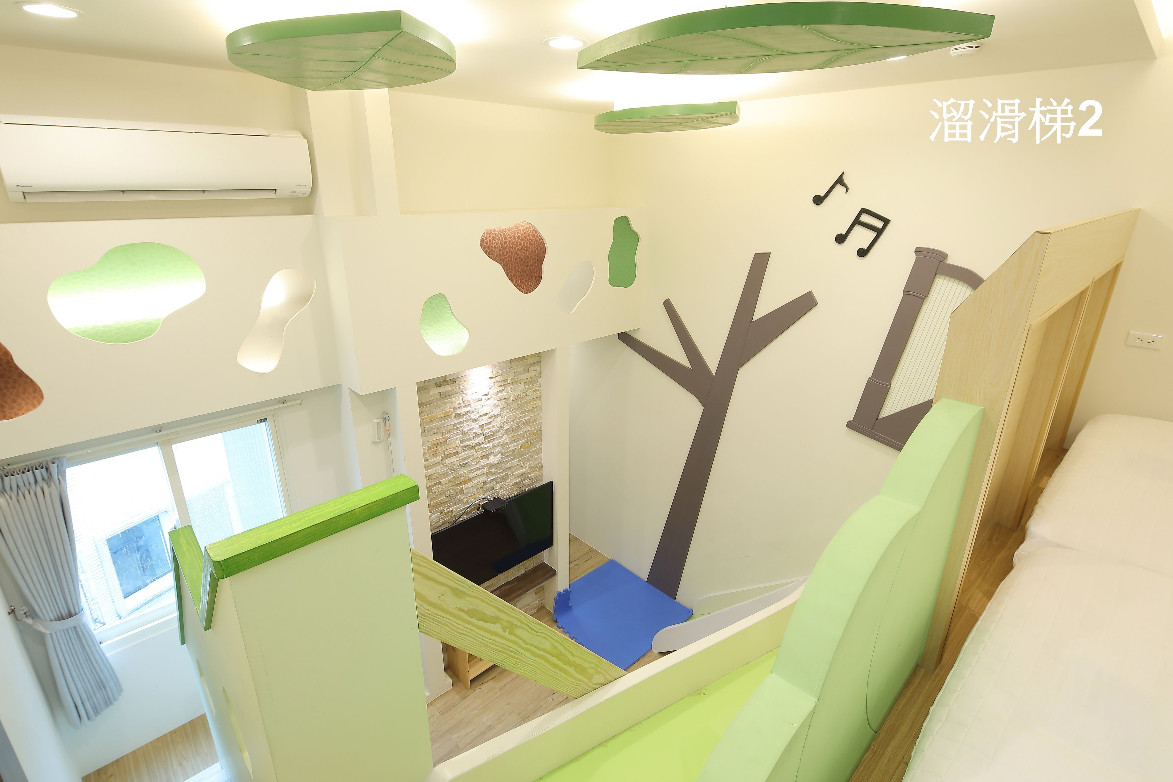 A room with a bed, a TV, and a window. There is a green wall with a tree design and a white wall with a window.