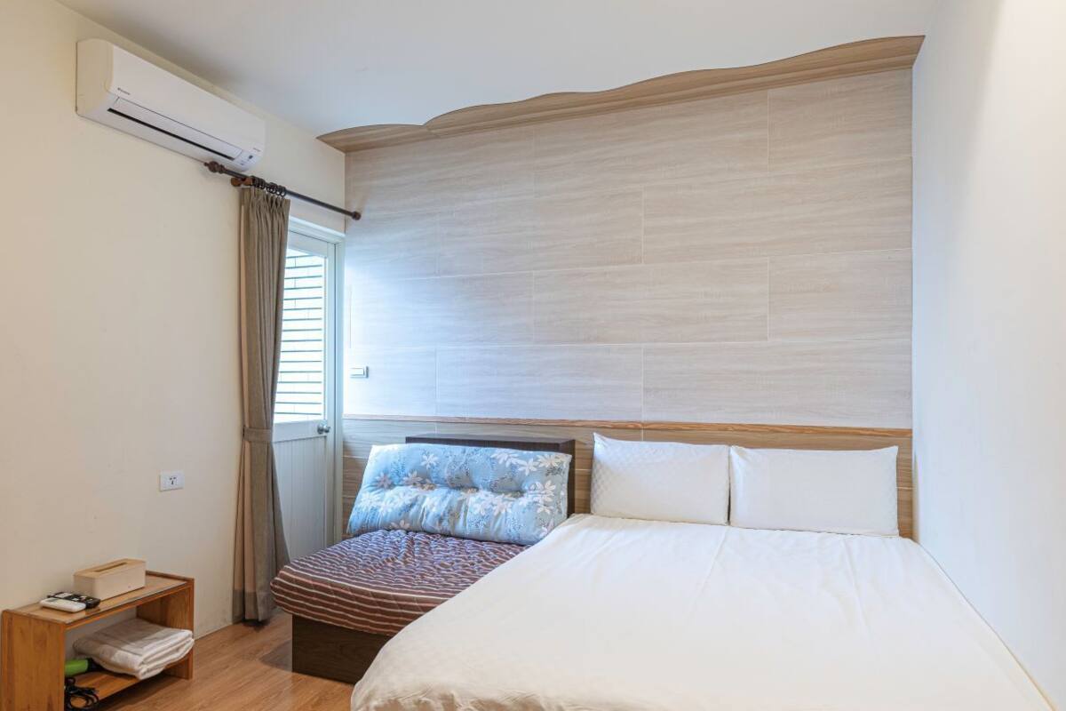 A bedroom with a large bed, a wooden nightstand, a window with curtains, and an air conditioning unit.