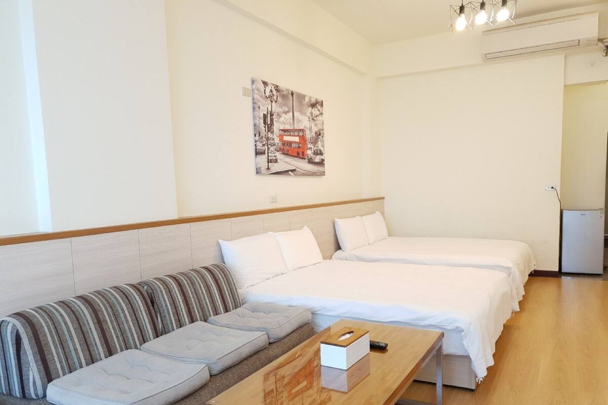 A hotel room with a sofa, two beds, a small table, and a wall-mounted air conditioner.
