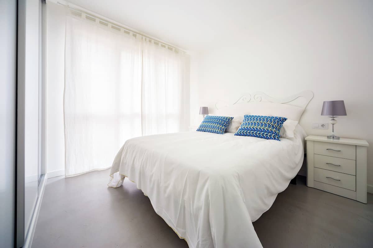 A bedroom with a bed, two bedside lamps, a nightstand, and a window with sheer curtains.