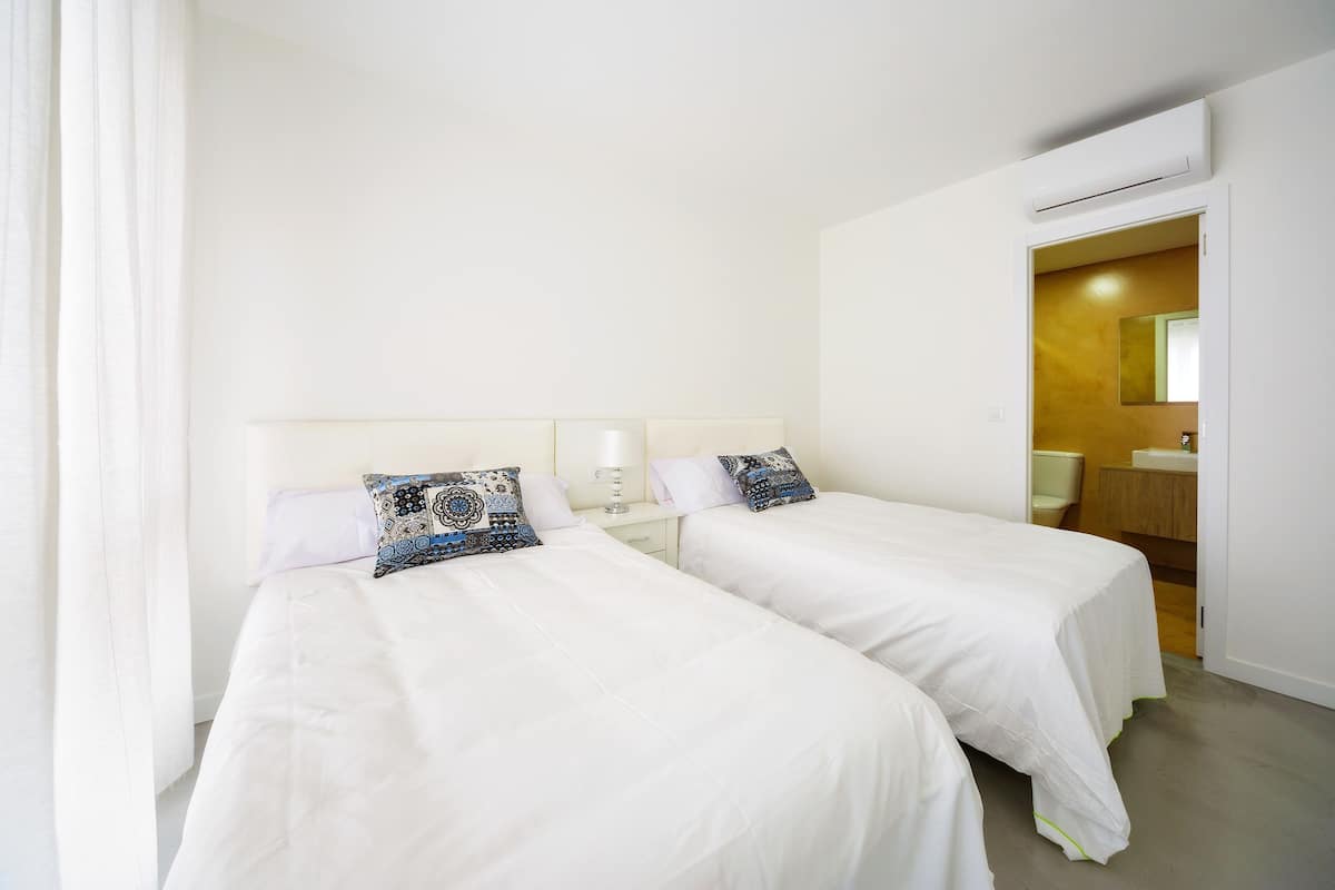 A hotel room with two beds, a nightstand, a lamp, and a bathroom visible through an open door.