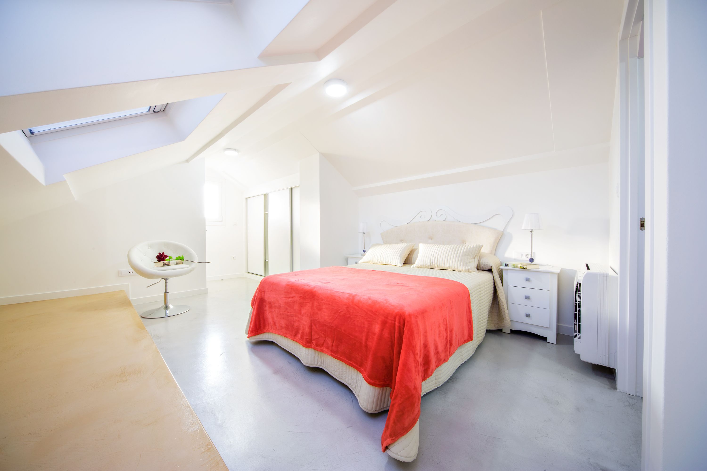A modern bedroom with a sloped ceiling, a bed with a red blanket, a white bedside table, and a chair with a vase of flowers.