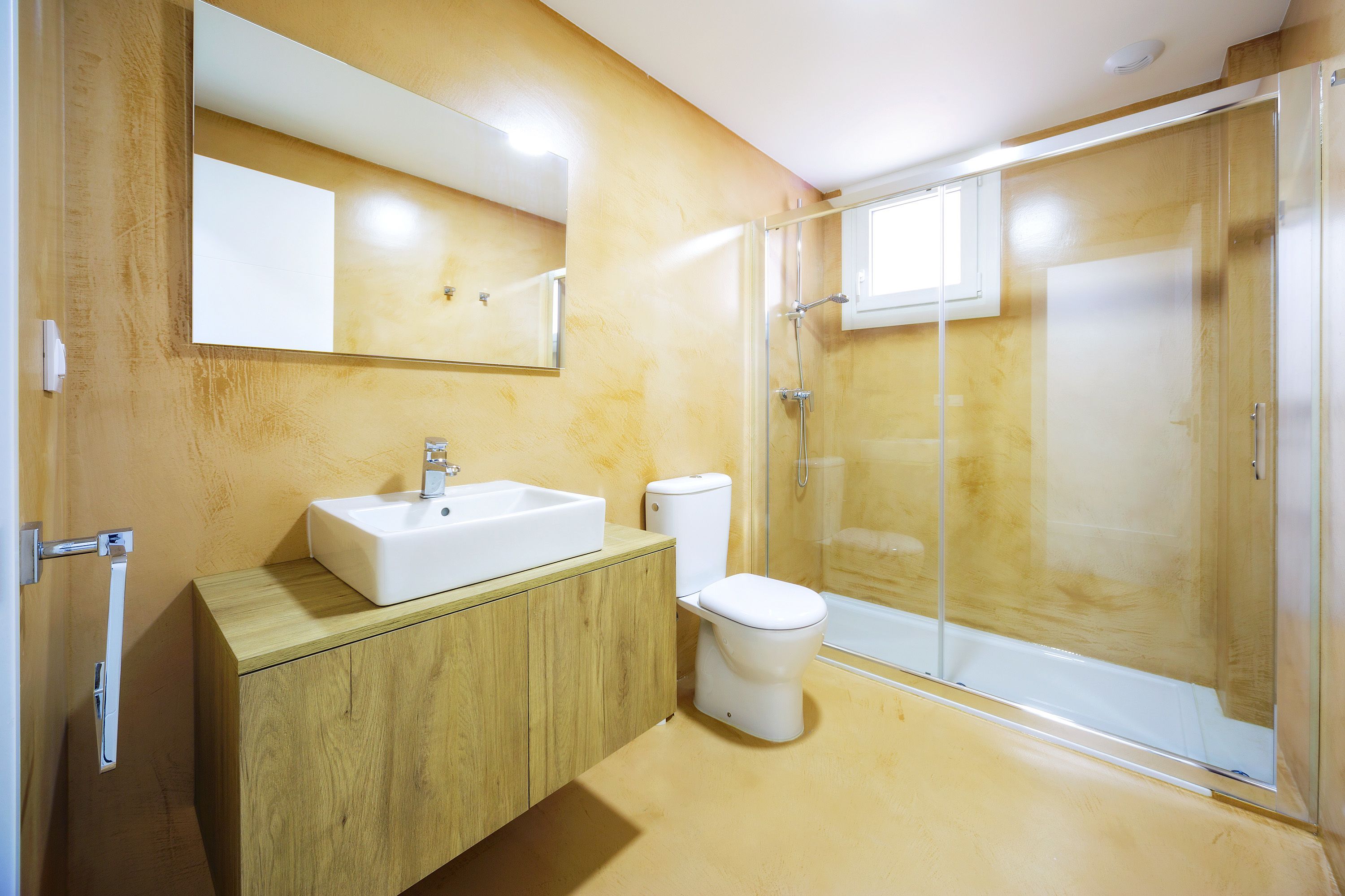 Un baño moderno con tocador de madera, un lavamanos blanco, un inodoro y una ducha con mampara de vidrio.