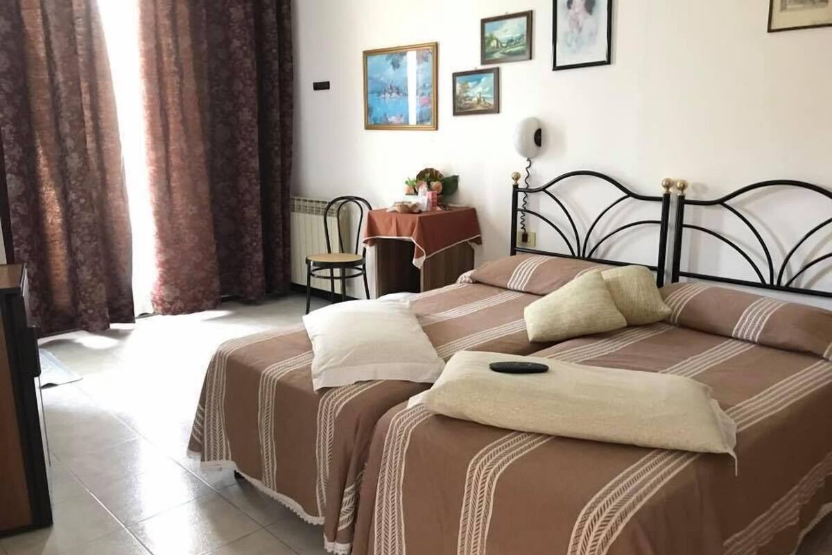 A bedroom with two beds, a chair, a table with a lamp, and framed pictures on the wall.