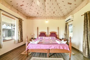 A neatly made bed with a pink bedspread and two pillows, a wooden headboard, a nightstand with a lamp, and a window with curtains. - Lohana Village Resort (Pushkar)