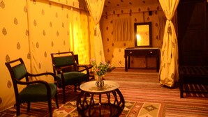 Una habitación con papel tapiz estampado, dos sillones tapizados de verde, una mesa redonda de madera con un jarrón de flores y una cama con colcha estampada.