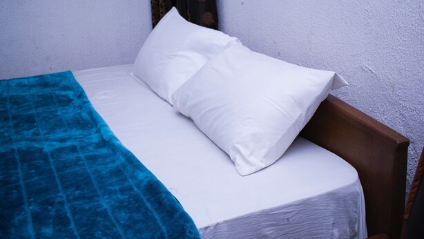 Un lit simple avec une literie blanche et une couverture bleue, une tête de lit en bois et un mur sobre agrémenté d’un rideau.