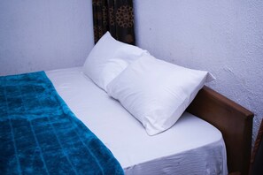 A single bed with white bedding and a blue blanket, a wooden headboard, and a plain wall with a curtain. - Day By Day Hotel (Lagos)