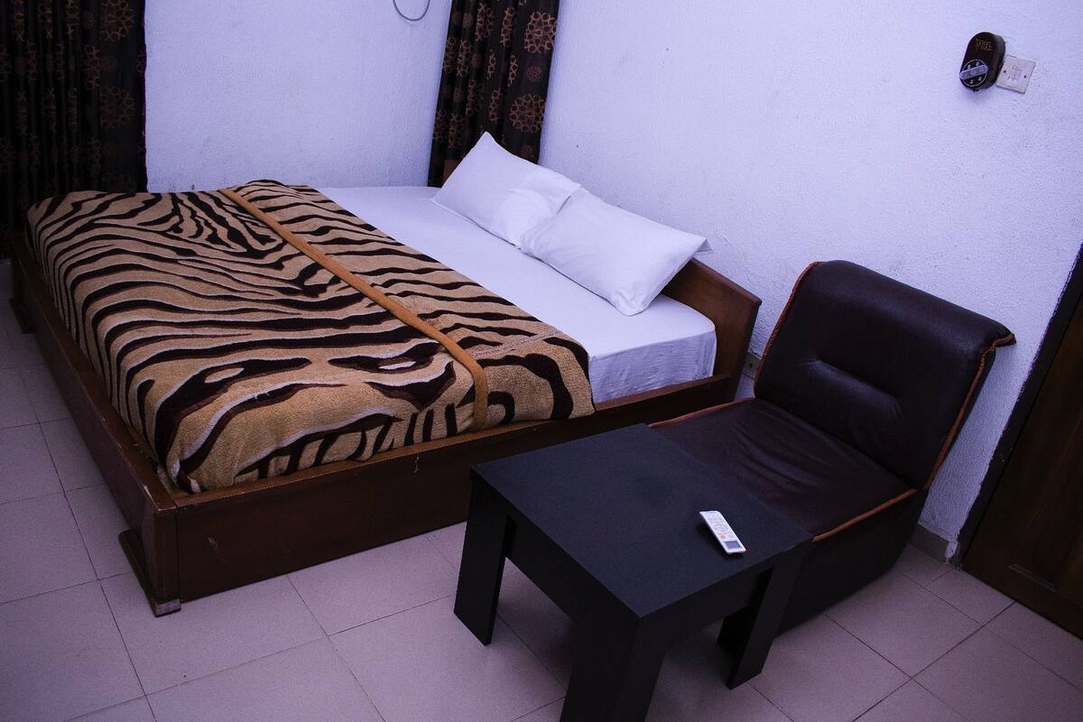 Una cama individual con una colcha de estampado de cebra, un almoháculo blanco y un cabecero de madera. Hay una mesita de noche pequeña de color negro con un control remoto encima. La habitación tiene una pared blanca y una cortina con diseño.