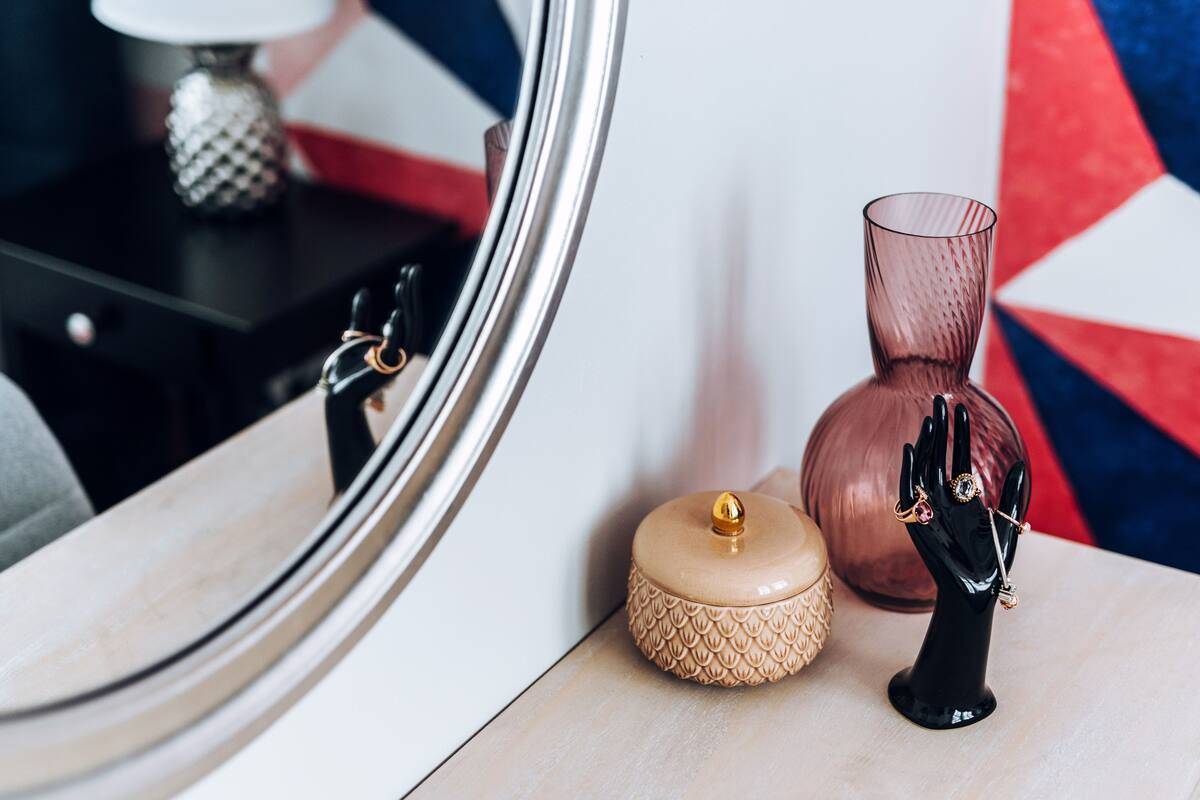 Un miroir reflétant une pièce dans laquelle on aperçoit une lampe, un vase et une sculpture décorative représentant une main, le tout posé sur une surface en bois.