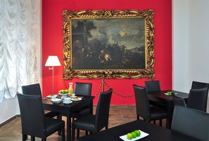 A dining area with a red wall, a large ornate gold-framed painting, black chairs, and a table set with plates, cups, and a pitcher of juice. - Hotel Lido (Reggio Calabria)
