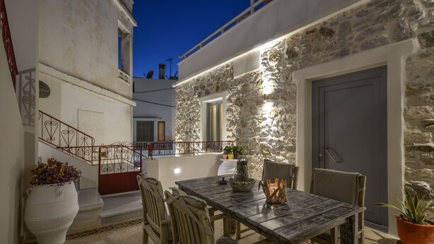 Un área de comedor al aire libre con paredes de piedra, una mesa, sillas, una planta en maceta y una escalera que lleva a un nivel superior.