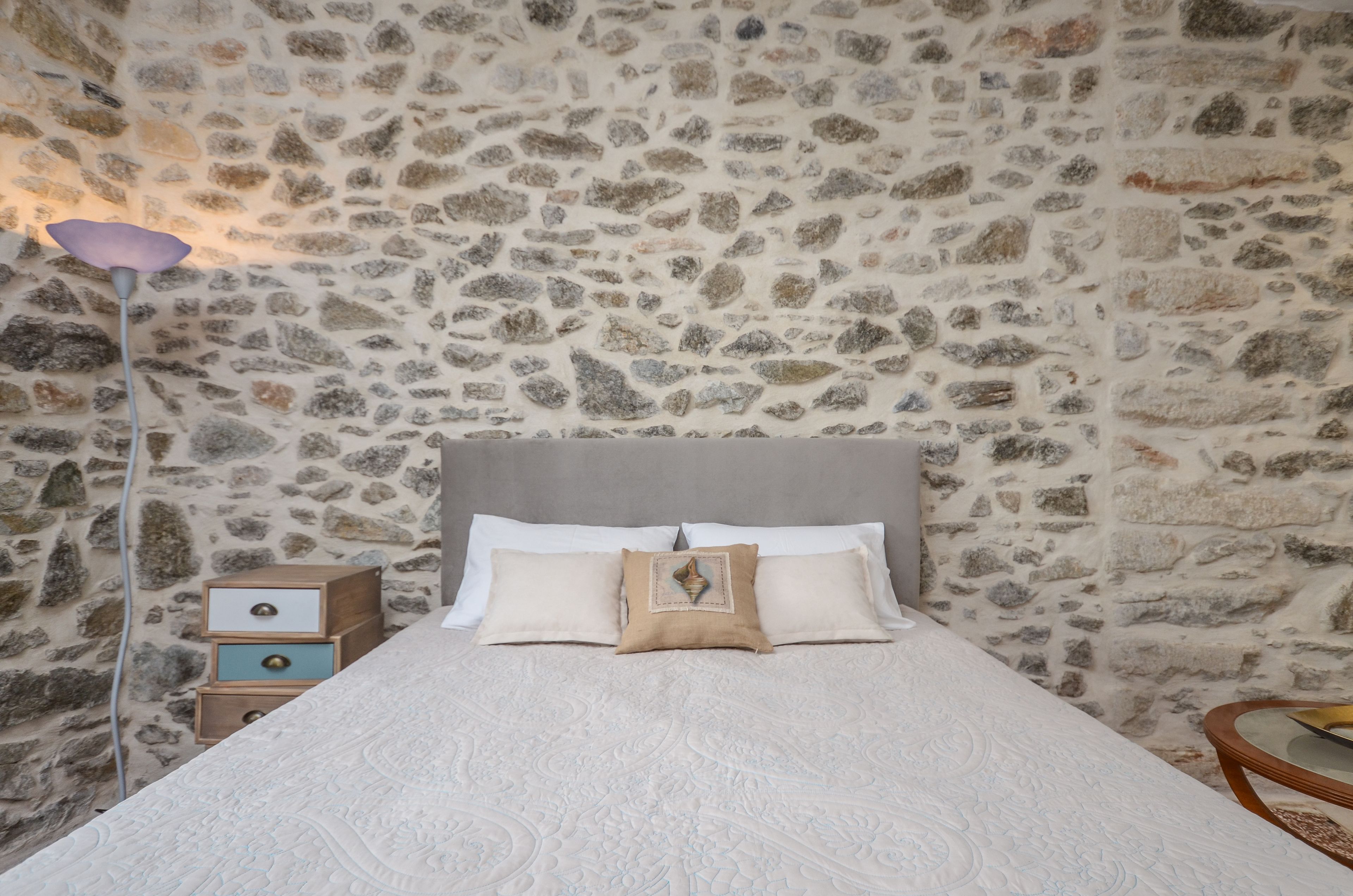 A bedroom with a stone wall, a bed with white bedding, a wooden nightstand, and a bedside lamp.