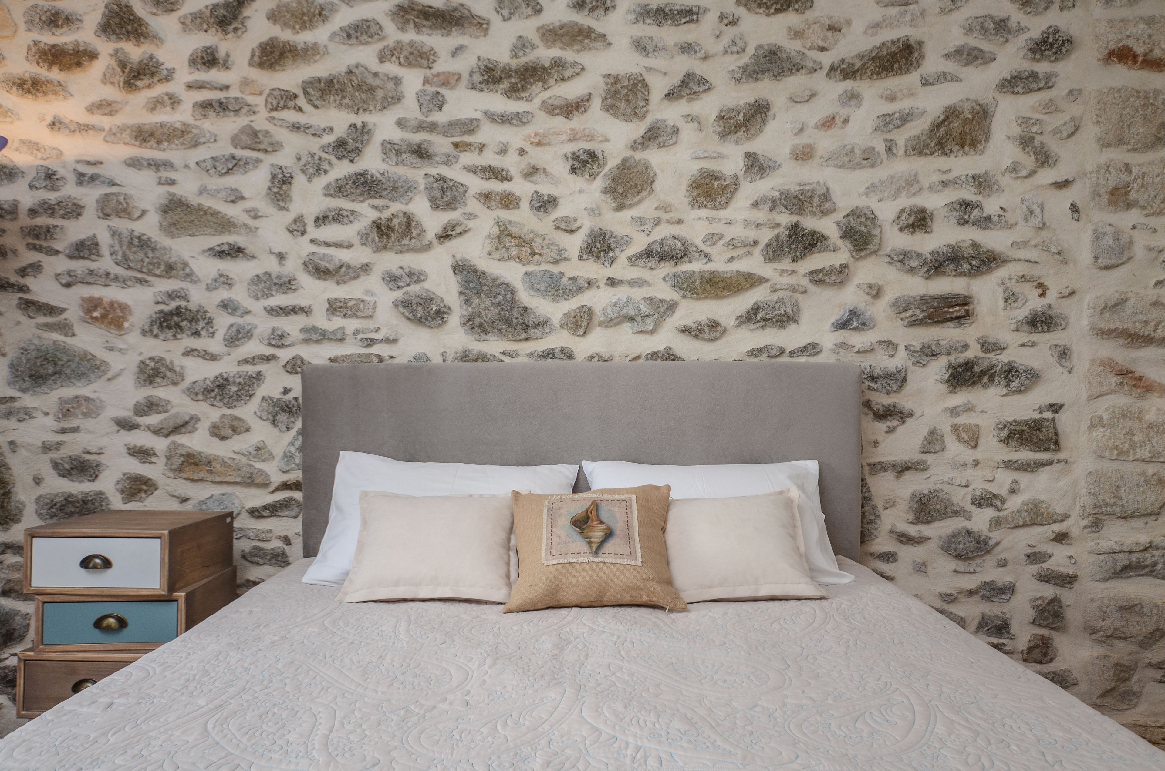 A bed with a stone wall background, a wooden nightstand with drawers, and a single decorative pillow.