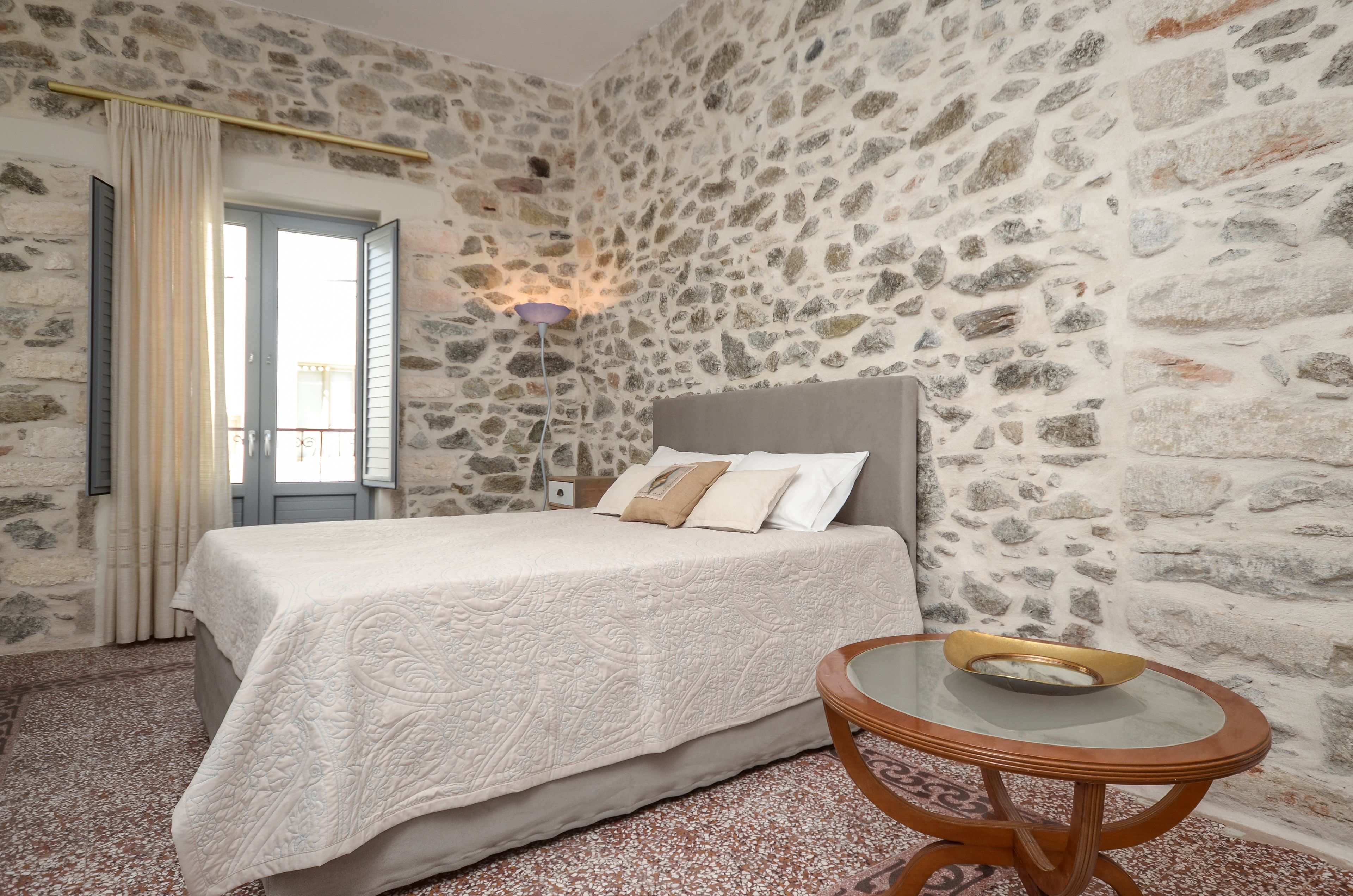 A bedroom with a stone wall, a bed with a white bedspread, a round wooden table with a bowl, and a window with curtains.