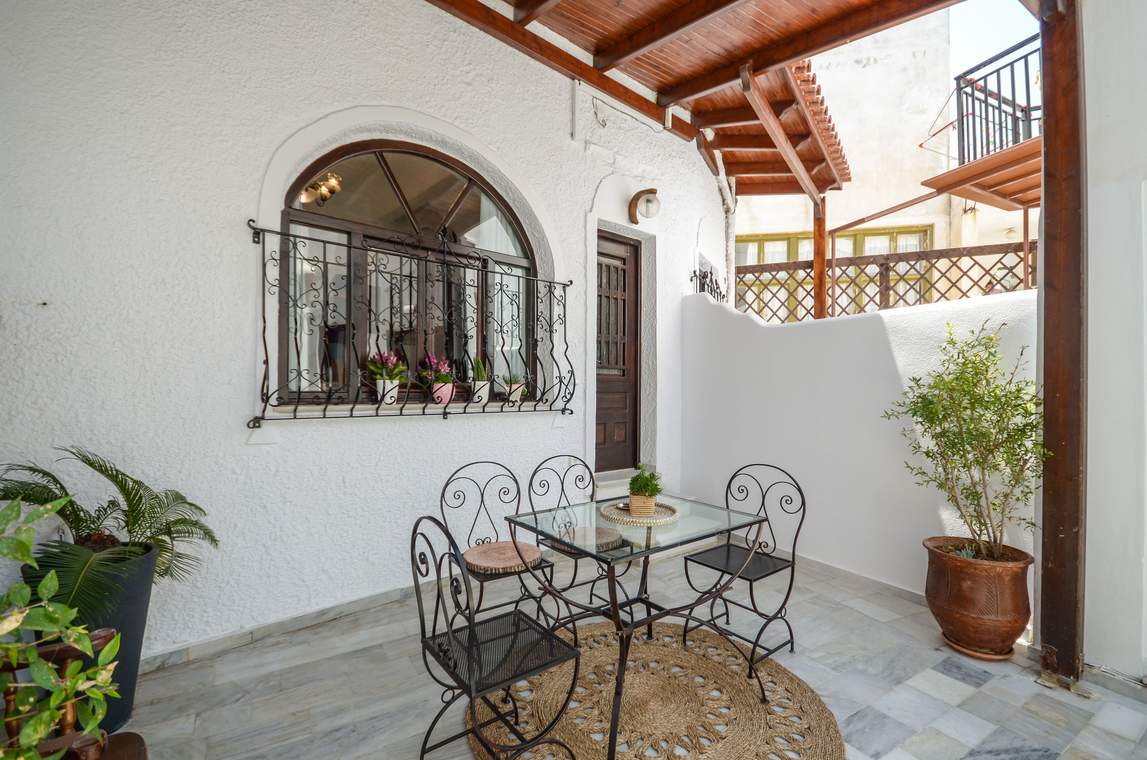 A patio with a glass table and metal chairs, surrounded by potted plants and a white wall with a window and door.