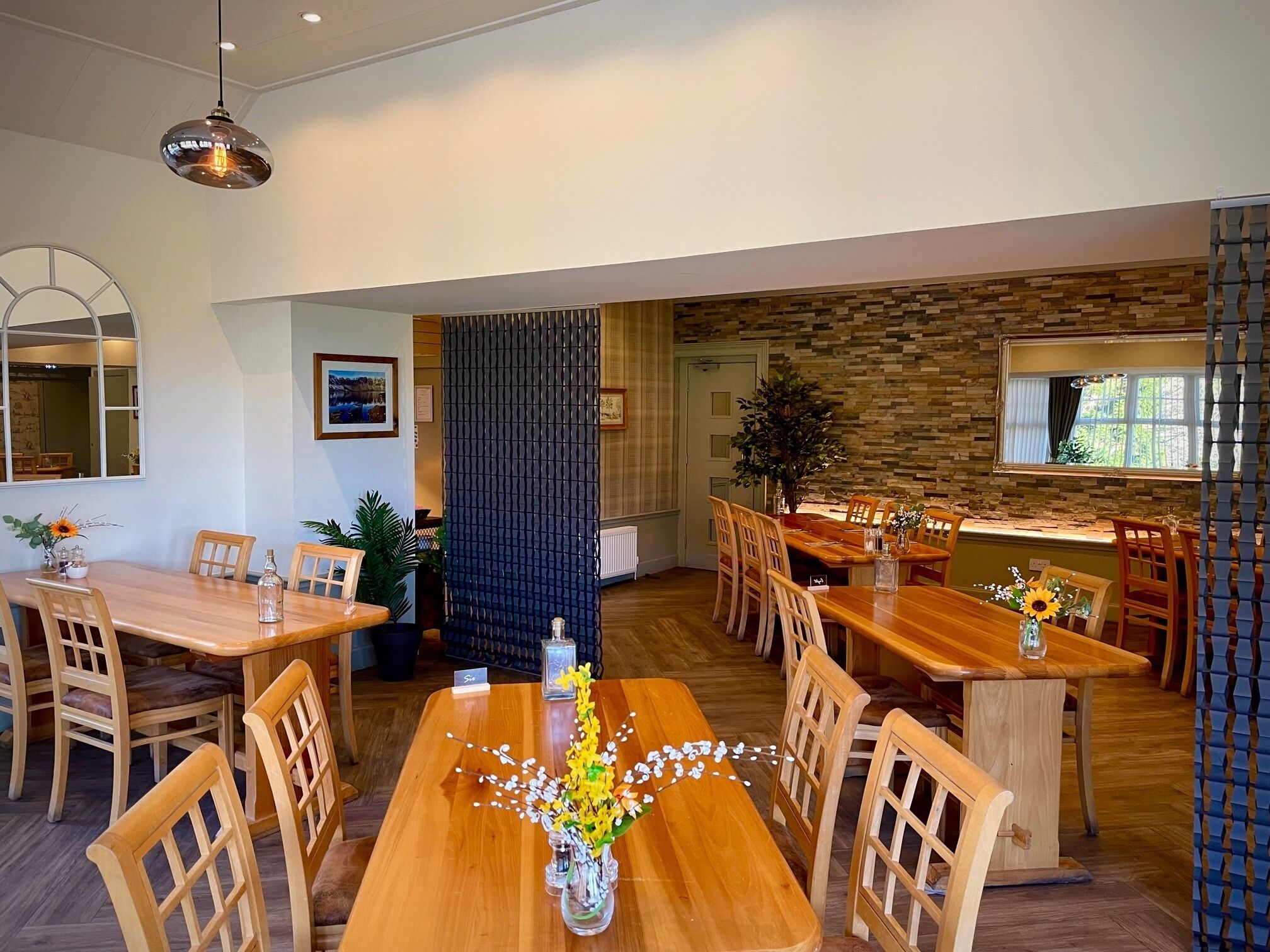 Un comedor con mesas y sillas de madera, un jarrón decorativo con flores y una pared de piedra al fondo.