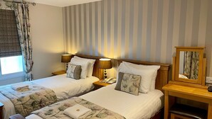 Habitación de hotel con dos camas, cada una con ropa de cama y almohadas estampadas con motivos florales, una mesita de noche de madera con lámpara, teléfono y espejo.