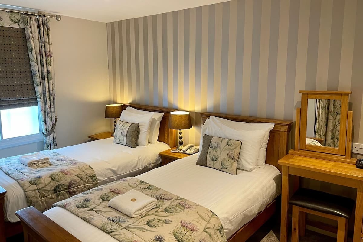 Habitación de hotel con dos camas, cada una con ropa de cama y almohadas estampadas con motivos florales, una mesita de noche de madera con lámpara, teléfono y espejo.