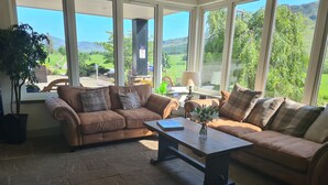 Una sala de estar acogedora con dos sofás marrones, una mesa de centro y vistas a un paisaje verde a través de amplios ventanales.