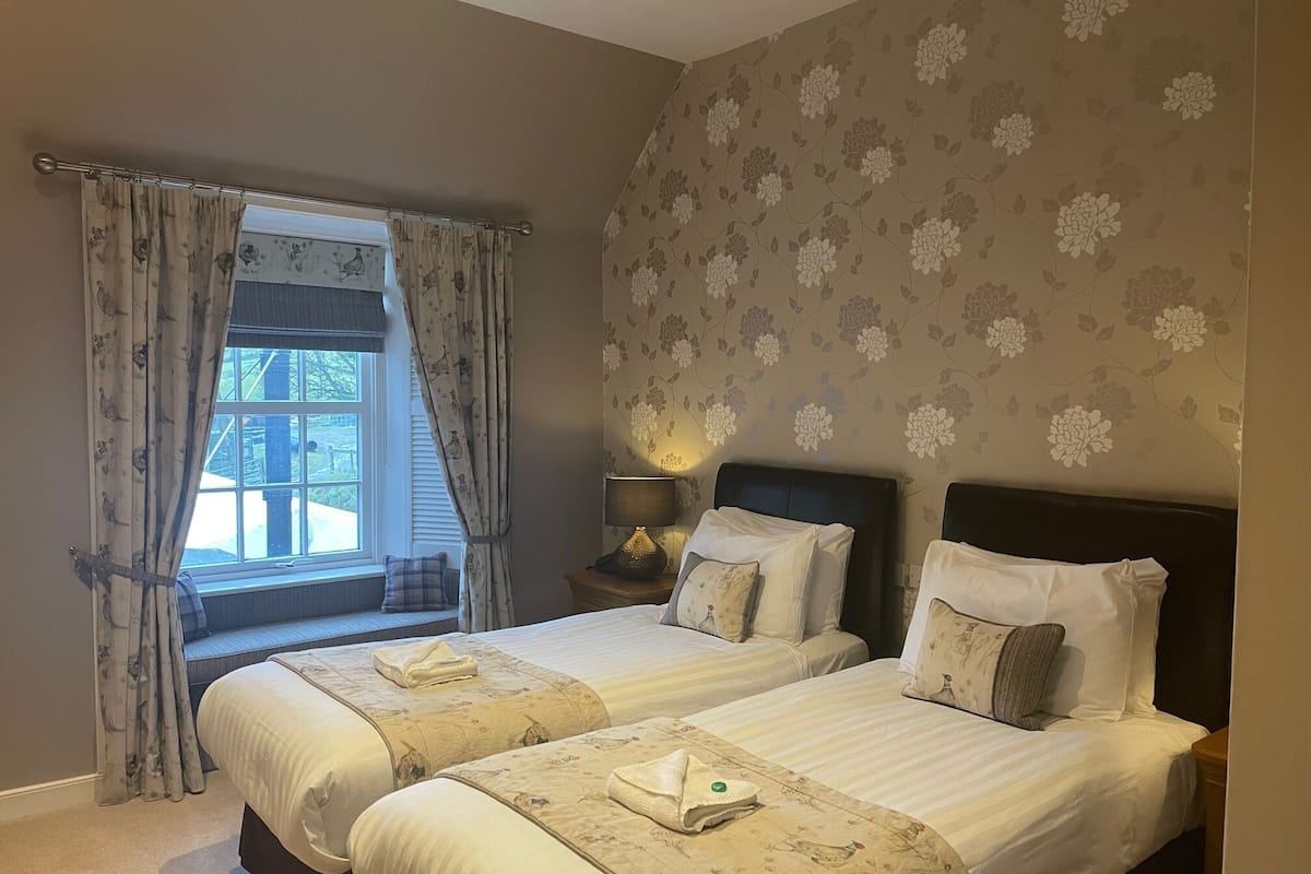 Un dormitorio con dos camas, papel tapiz floral, una ventana con cortinas y una lámpara de noche.