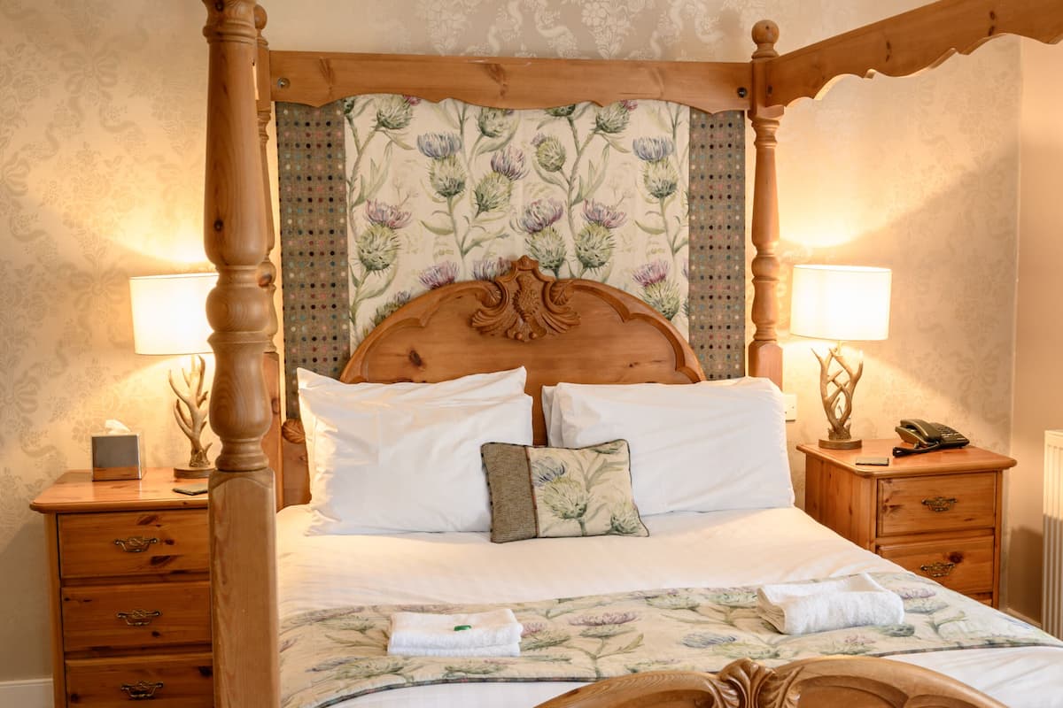 Una cama con dosel de madera, un cabecero floral, dos mesitas de noche con lámparas y una mesita auxiliar con teléfono.