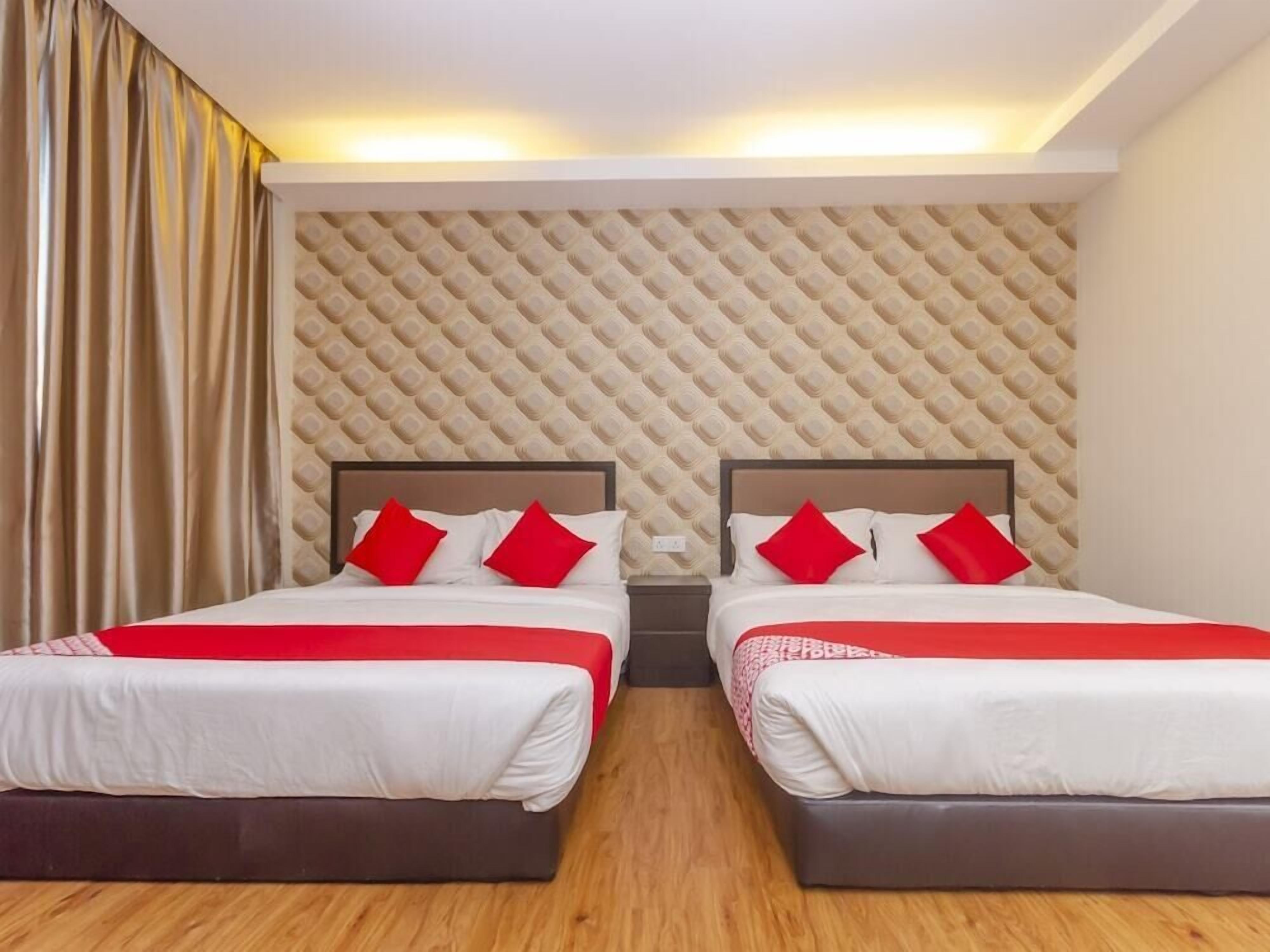 A hotel room with two beds, each with a headboard, red pillows, and a red and white bedspread. The room has a patterned wall and a wooden floor.