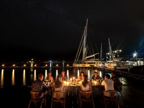 Un groupe de personnes dînant à une table en plein air, près d’un port de plaisance, la nuit.