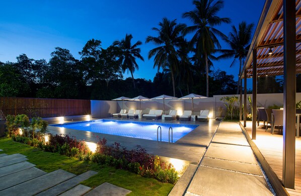 A swimming pool with lounge chairs and umbrellas, surrounded by palm trees and a wooden deck.