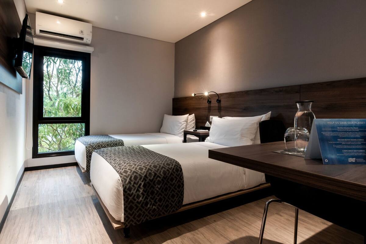 Una habitación de hotel moderna con dos camas, un escritorio, una mesita de noche, un ventanal grande y un equipo de aire acondicionado.