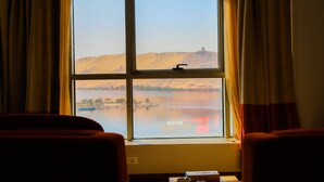 Una habitación con un ventanal grande que da a un lago y colinas. Hay una cama y una mesa con una caja y una taza encima.