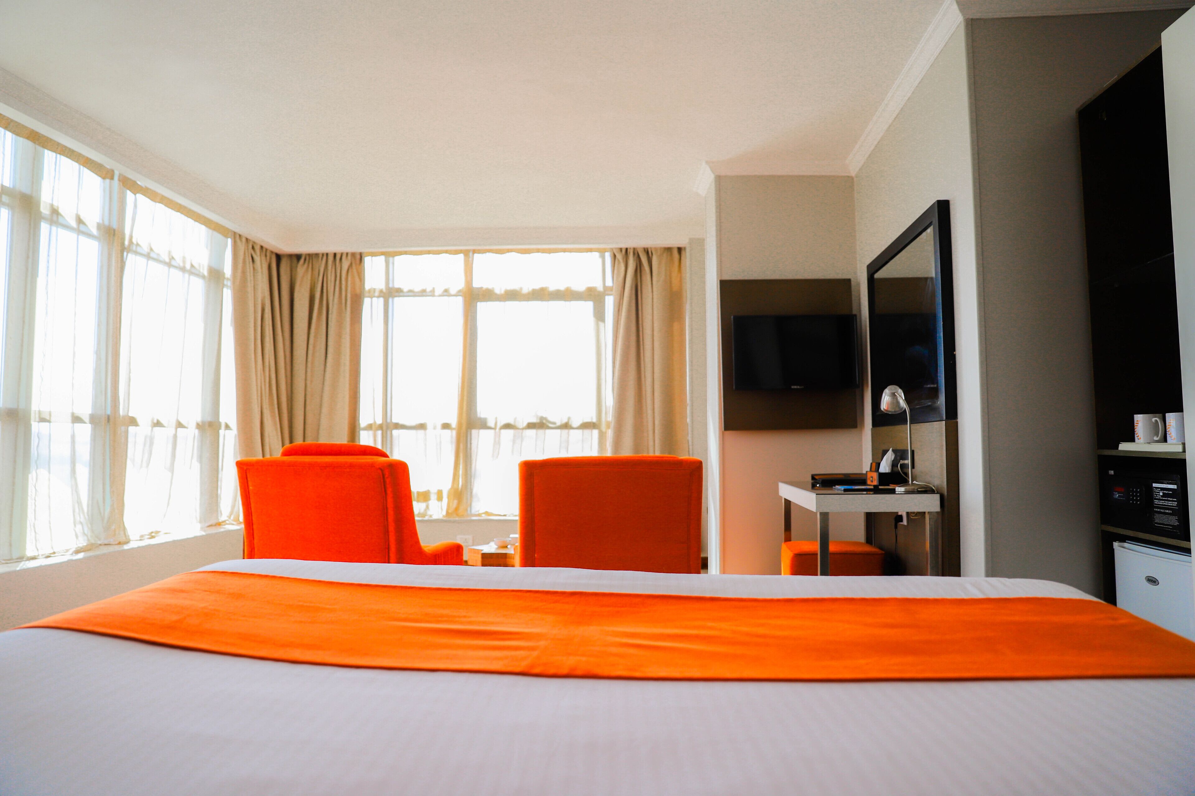 A hotel room with a large bed, two orange chairs, a desk with a lamp, a television, and a minibar.