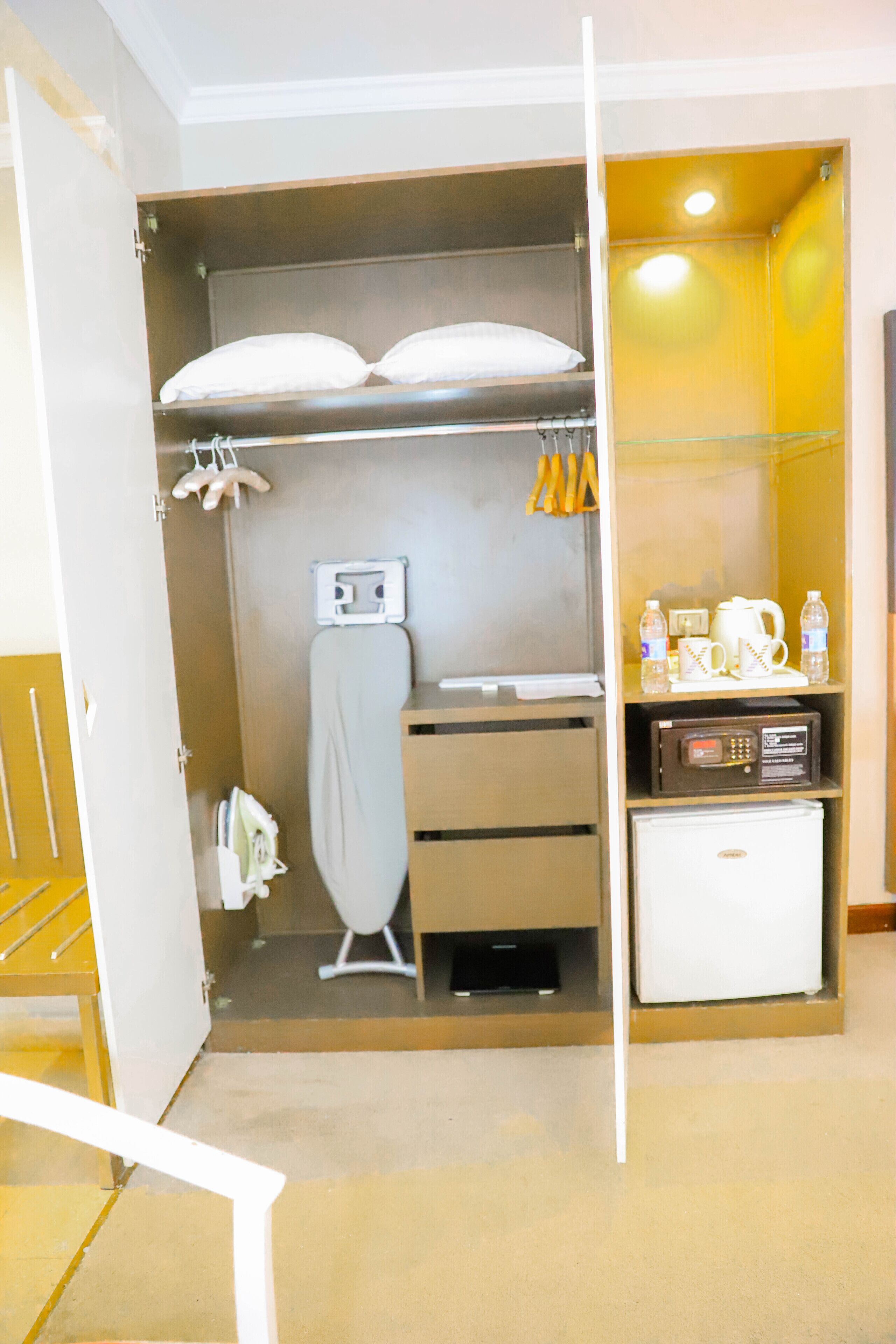 A compact hotel room with a wardrobe, a small refrigerator, a microwave, and a shelf with toiletries.