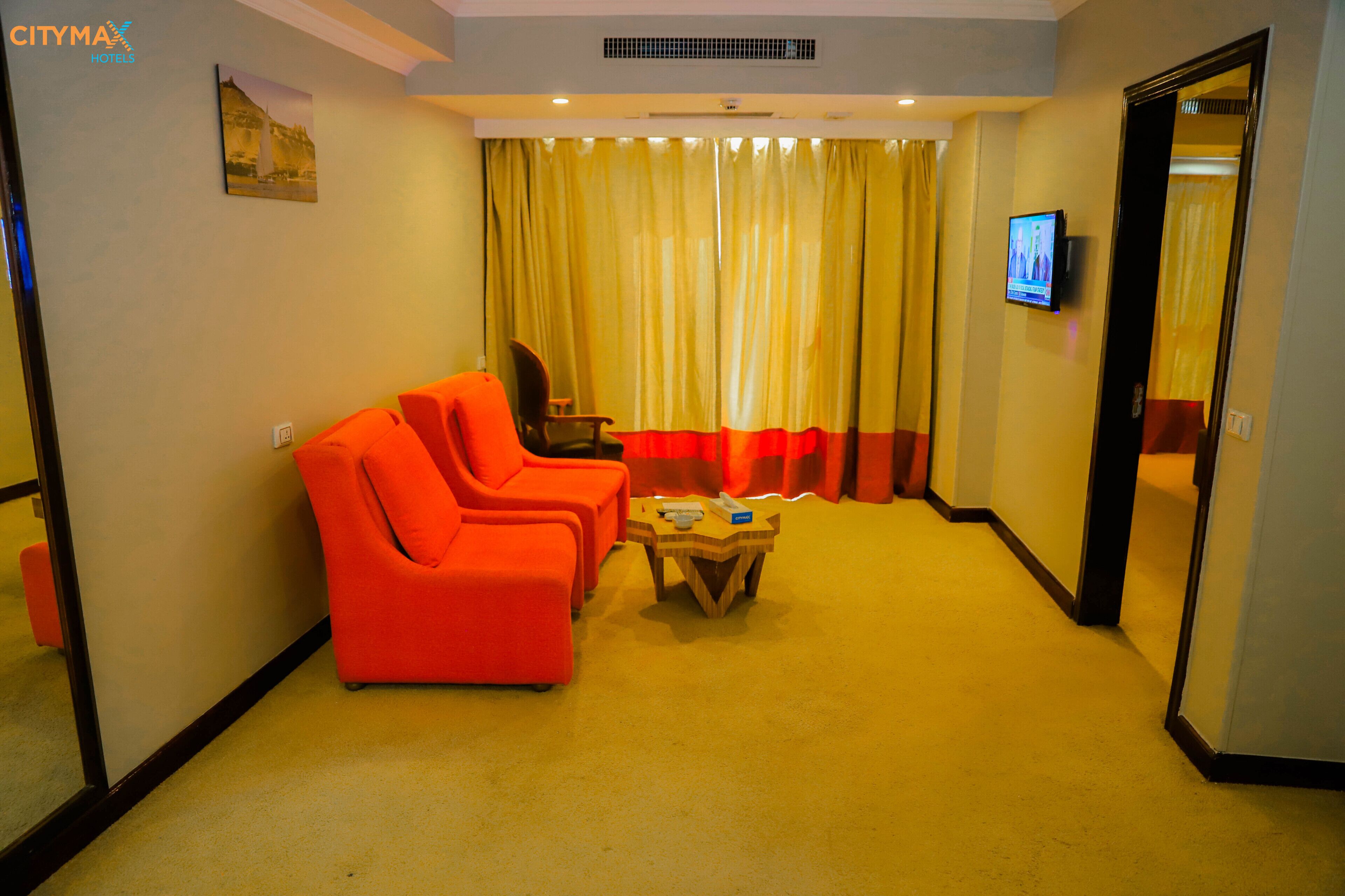 A hotel room with a seating area featuring two orange chairs, a small wooden table, and a television on the wall.