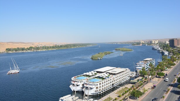 Un río ancho con un barco crucero atracado a la derecha, un velero en el agua y un paisaje urbano con edificios y vegetación a lo largo de la orilla.