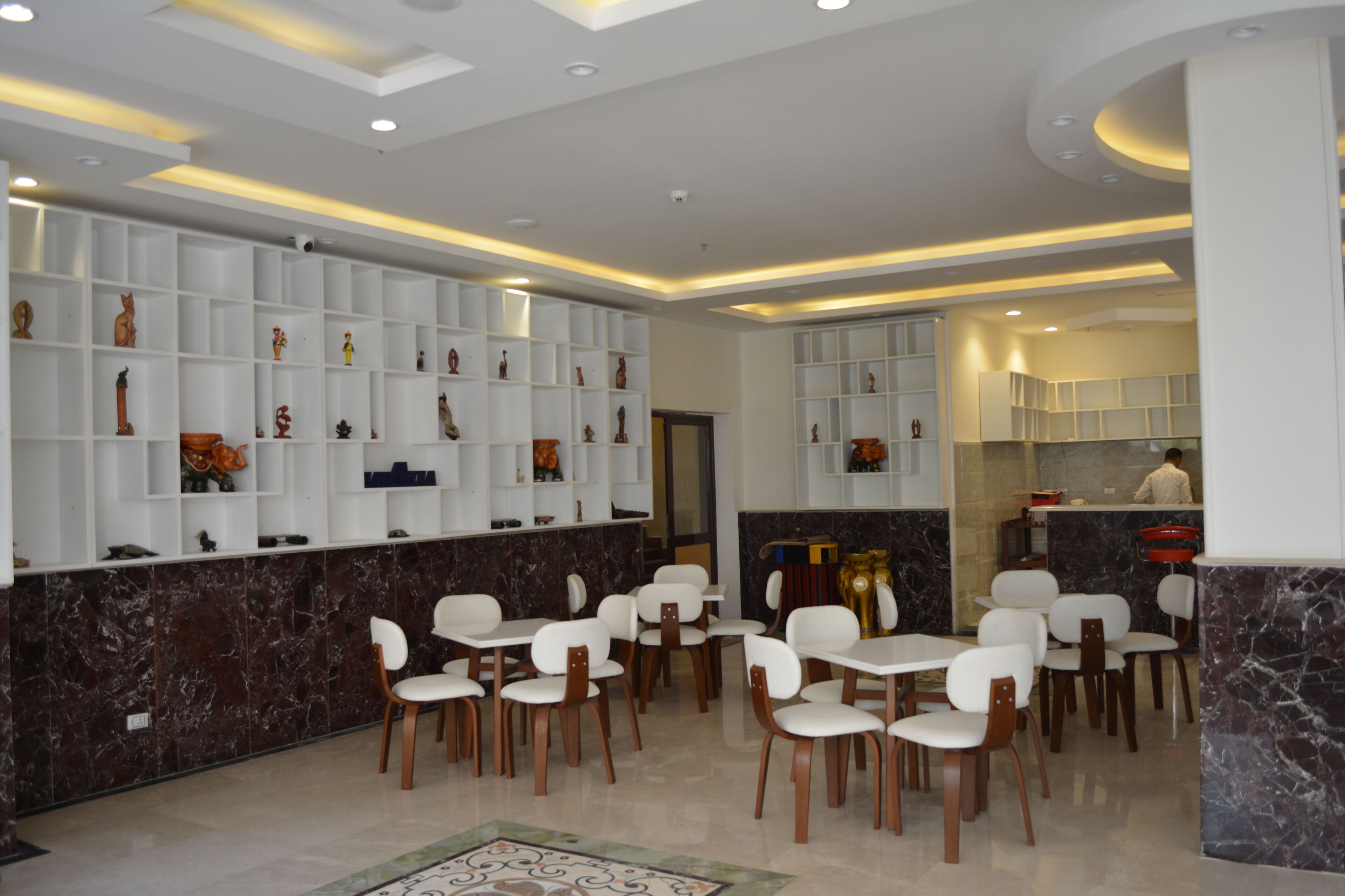 A modern dining area with white chairs and tables, marble walls, and shelves with decorative items.