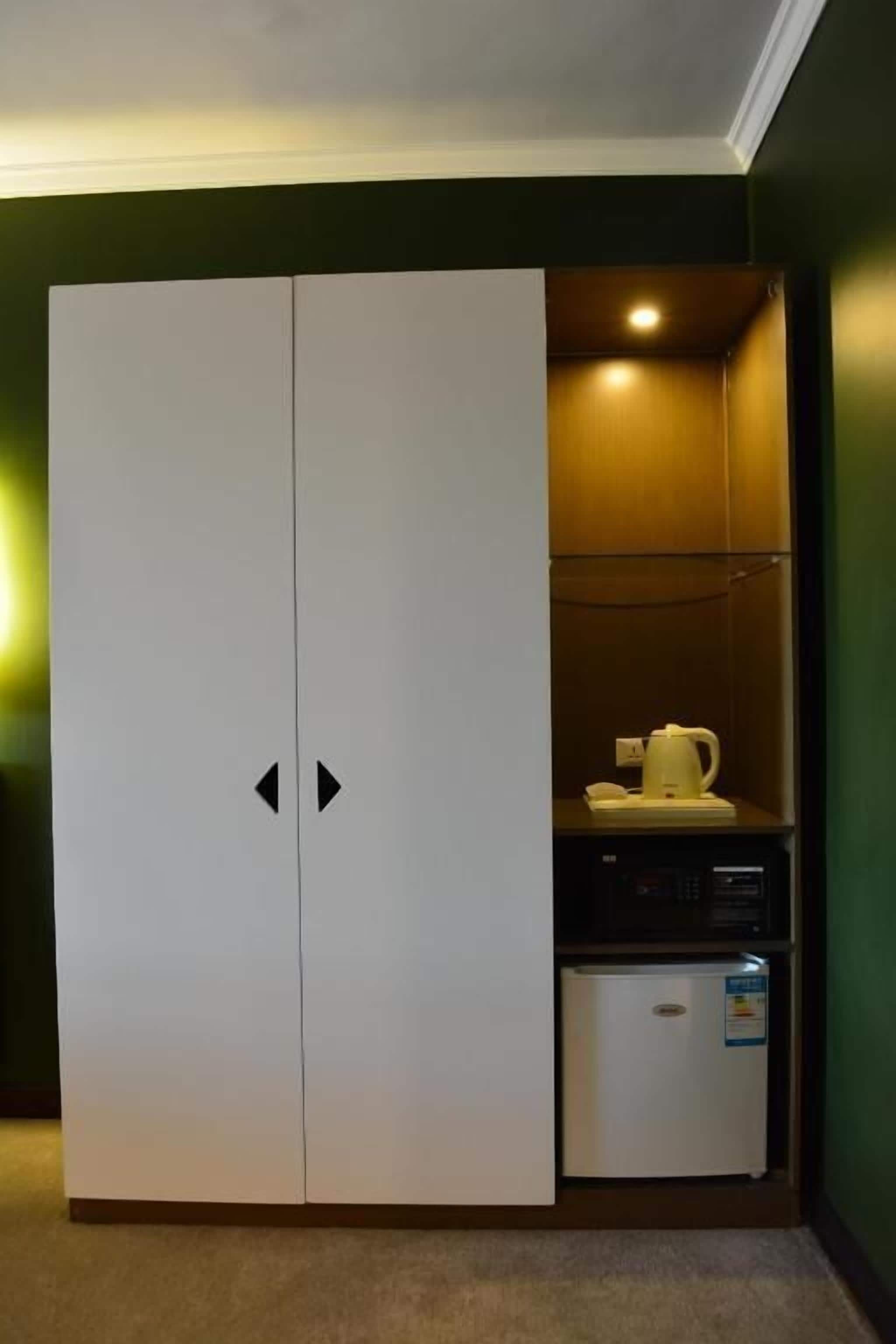 Una habitación con un armario blanco, una kitchenette pequeña con hervidor y microondas, y una pared verde.