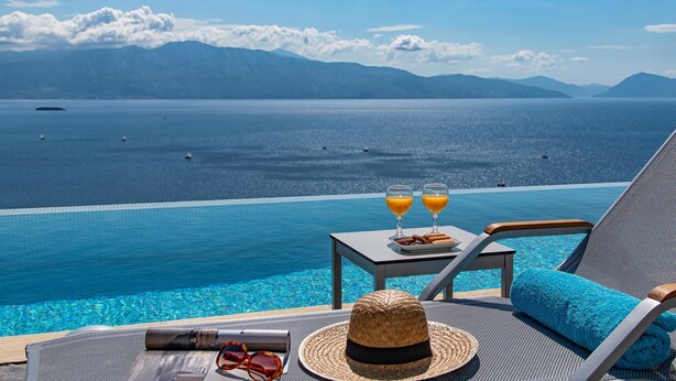 A poolside view with a clear blue sea, mountains in the background, and a table with glasses of orange juice and a hat. - Ionian Heaven Villas (Lefkada)